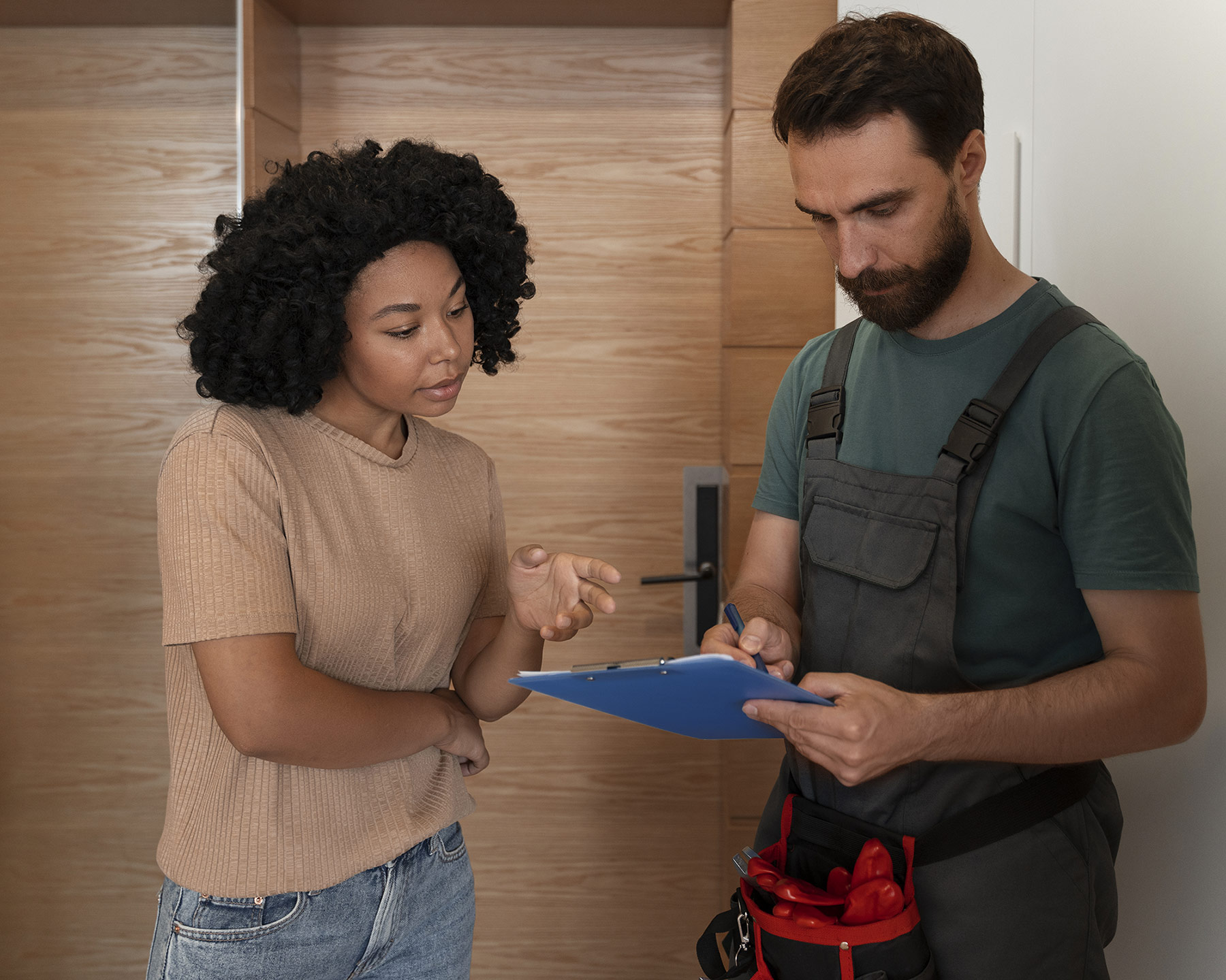 Stock photo of a man and woman standing indoors. The man is wearing overalls with a toolbelt, and is looking intently at a checklist he is holding. The woman is also staring down at the checklist, and is gesticulating like she has something to contribute.